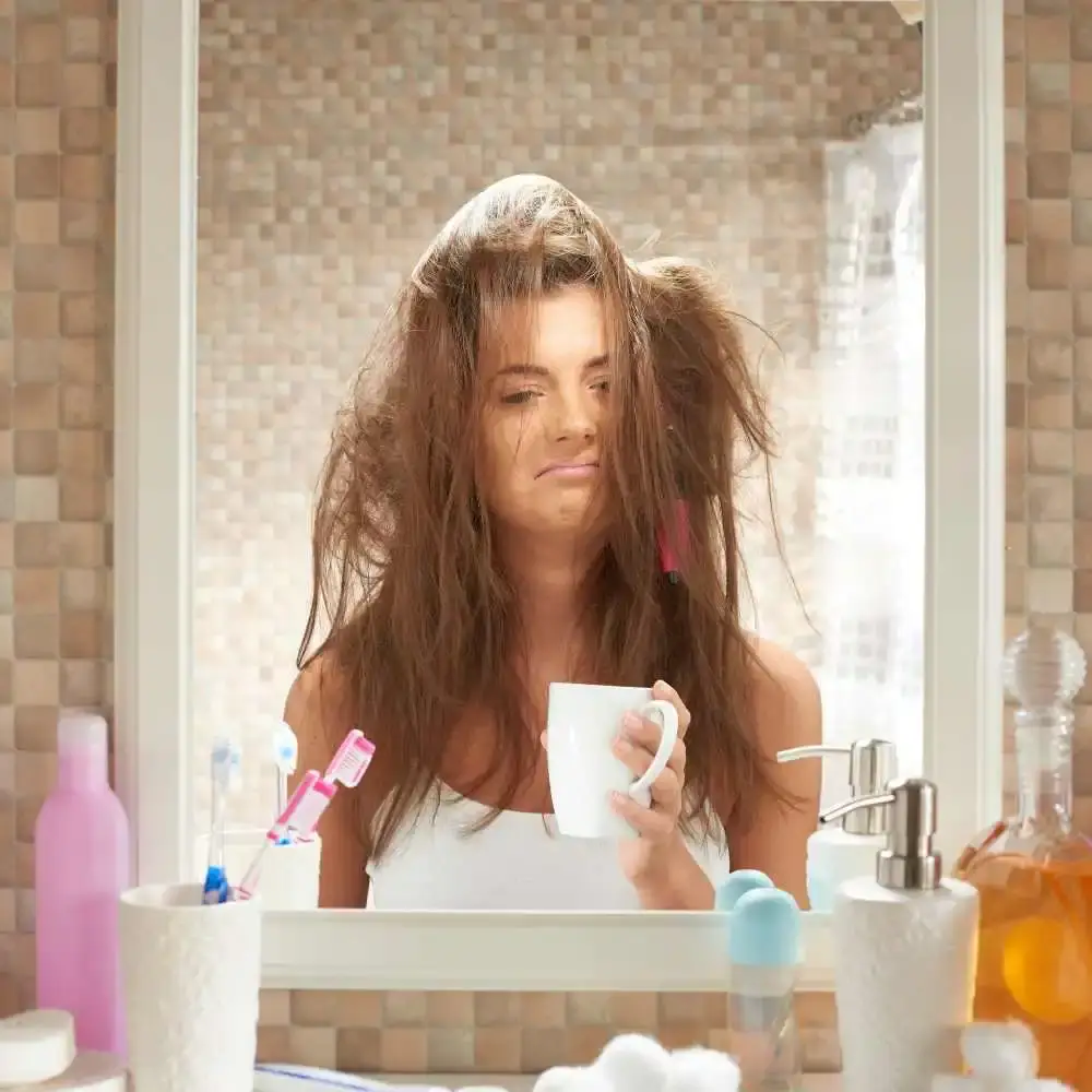 young woman with tangled hair frowning in front of the mirror holding a mug