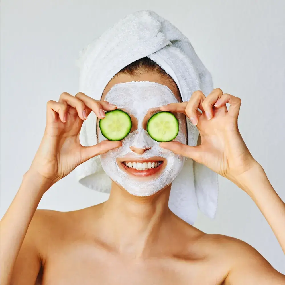 woman smiling with face mask and holding two slices of cucumber